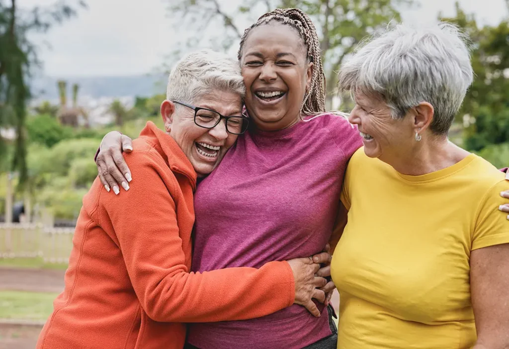 Group of three cheerful women embracing and laughing together in a park, dressed in colorful casual clothes.