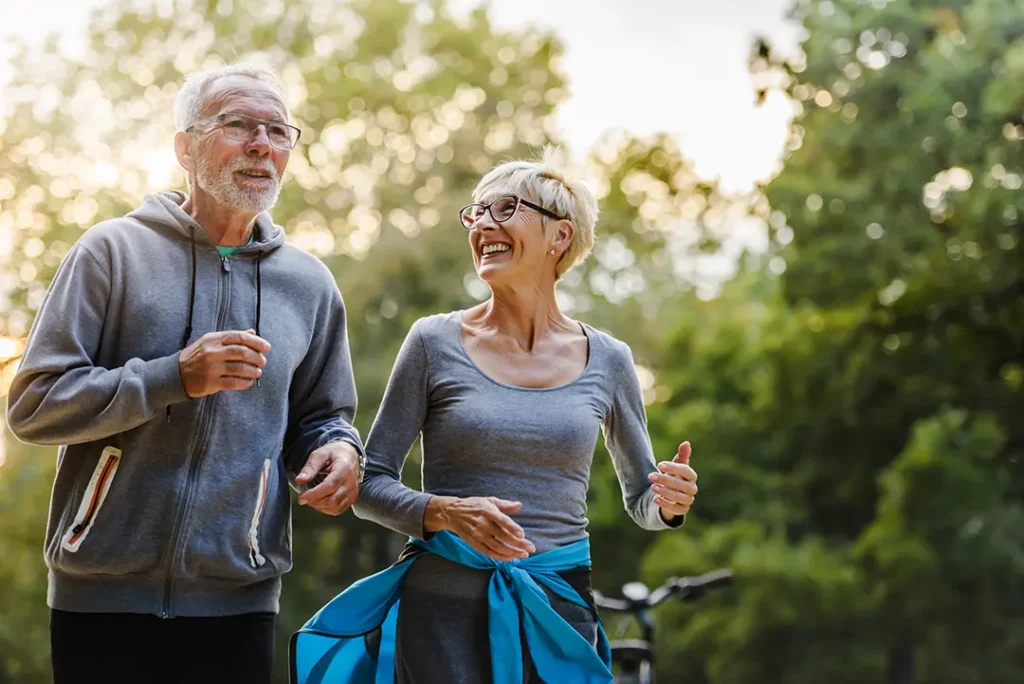 An older couple jogging together outdoors, smiling and enjoying each other's company with trees and sunlight in the background.