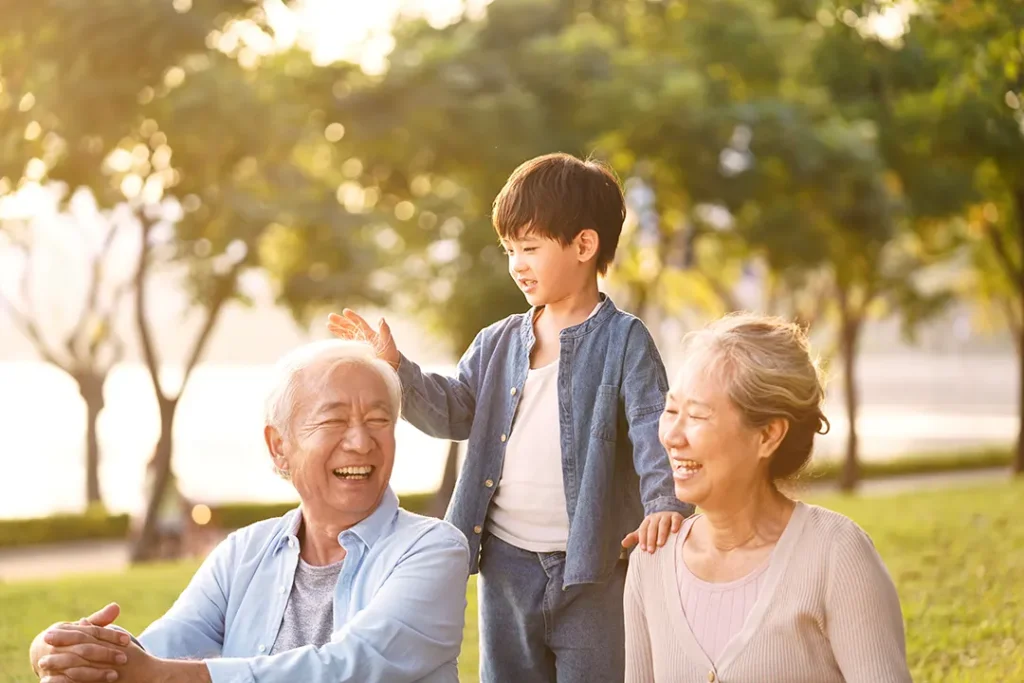 Grandparents laugh joyfully as their grandson stands behind them in a park, resting his hands affectionately on their shoulders in the golden sunlight.