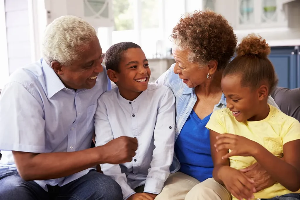 Two grandparents sitting on a couch, laughing with their smiling grandson and granddaughter in a bright, cozy home.
