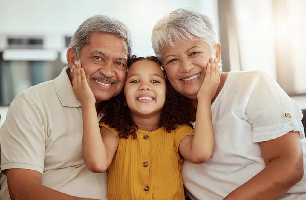 Smiling grandparents sit closely on either side of their granddaughter, who is holding both of their faces in her hands with joy.