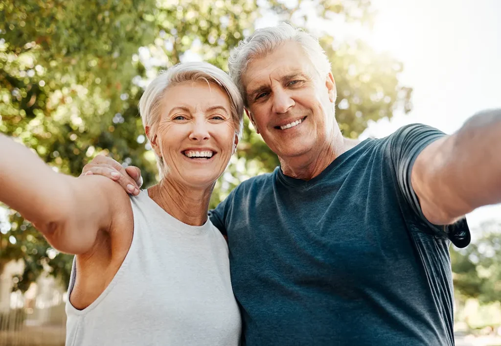 Smiling older couple taking a selfie outdoors, standing close together with arms extended toward the camera on a sunny day.