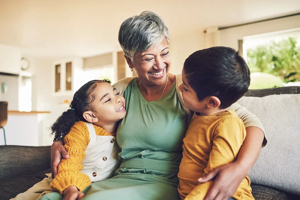An older woman sits on a couch, smiling joyfully while hugging two young children who lean into her with big grins in a cozy, sunlit living room.
