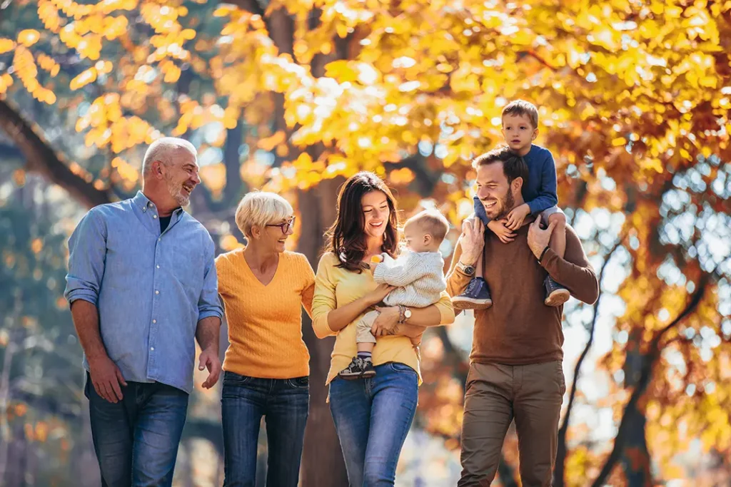Three generations of a smiling family walk together through a sunlit park with golden autumn leaves, including grandparents, parents, and two young children, one held in arms and the other riding on shoulders.