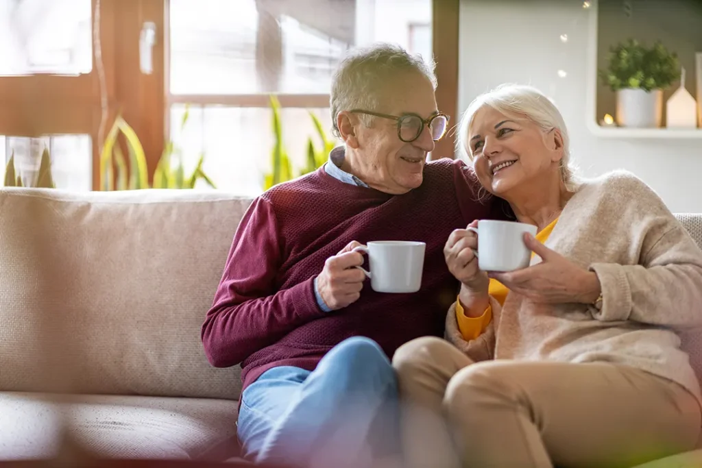 An older couple sits close together on a couch, smiling warmly at each other while holding white mugs, in a bright and cozy living room.